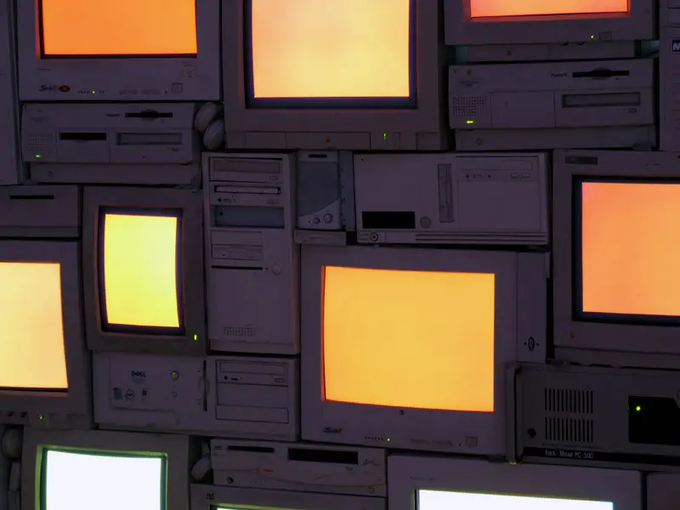 A wall of stacked old computer monitors and towers displaying solid orange and white screens, illuminated in a dim setting.