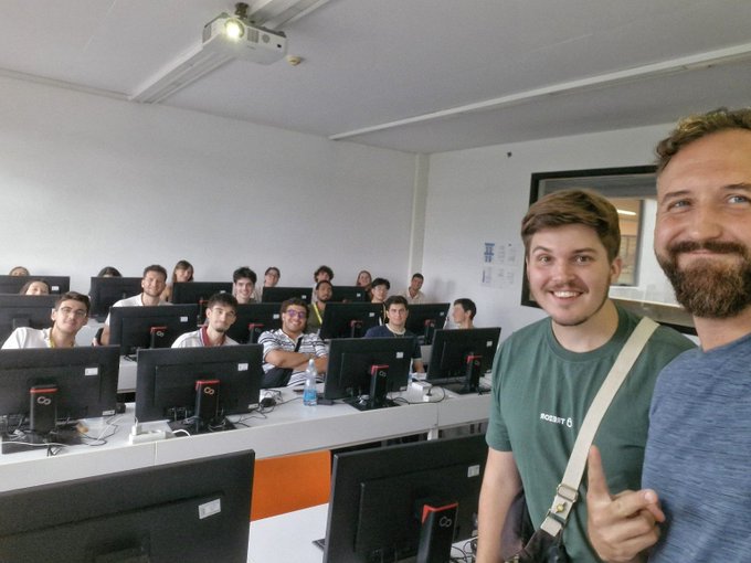 Two smiling men posing for a selfie in a computer lab filled with students seated at rows of monitors under a ceiling-mounted projector.