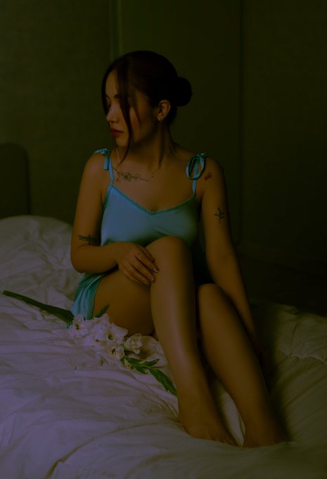 Moody, dimly-lit portrait of a woman in a light blue/teal slip dress sitting on a bed with white sheets and flowers, with visible tattoos on her arms.