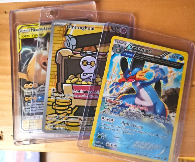 Three Pokémon trading cards in clear protective sleeves rest on a wooden surface, featuring Pikachu & Zekrom (Tag Team), Gimmighoul, and Swampert.
