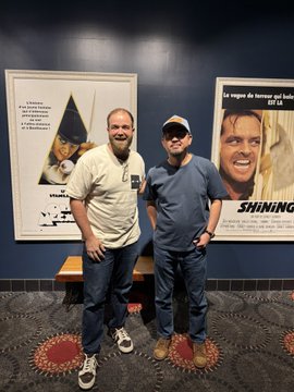 Two men stand together smiling in a movie theater lobby in front of posters for "The Shining," with Spanish text "La niego de horror qué bebé" and "COLORES" visible.