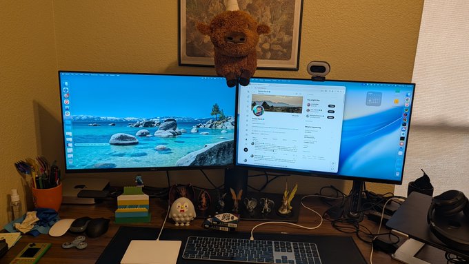 A newly set up desk at Sei Labs features a dual-monitor setup with a Windows desktop background displaying a scenic lake landscape, while the second monitor shows what appears to be a browser or application interface with some text content. The workspace is personalized with a teddy bear perched on top of the monitors, framed artwork on the wall, and various small items including figurines, a keyboard, mouse, and office supplies arranged on the wooden desk.