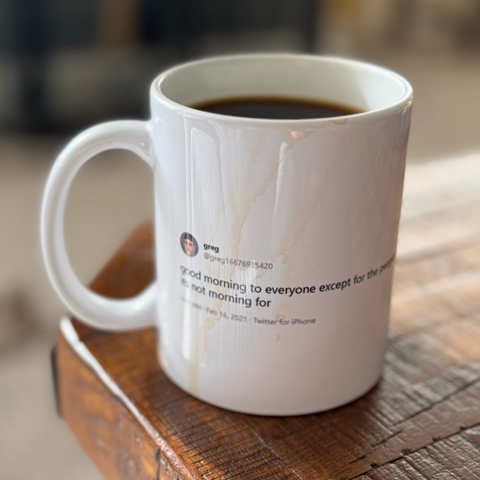 A white coffee mug filled with coffee sits on a wooden surface, featuring a printed tweet from user "greg (@greg1667693420)" dated "Feb 16, 2021" that reads "good morning to everyone except for the people who are not morning for." The image playfully connects to the post's cheerful Friday morning vibe, with the tweet's humorous "morning person" jab contrasting the optimistic start to the day.