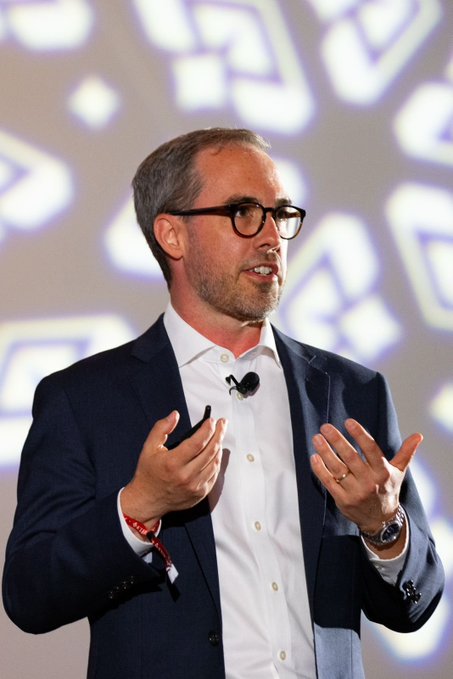 The image features a man wearing glasses, a suit jacket, and a white shirt standing in front of a screen with a patterned background. The man appears to be giving a speech or presentation, as he has his hands outstretched while speaking. The image also contains text that reads "AI'S PROMISE depends on it" and "@filFoundation". This suggests that the man might be discussing the importance of AI in today's world, particularly in relation to data storage and privacy concerns.