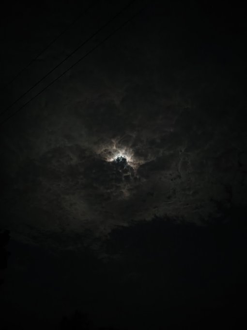 A nighttime sky photo showing the moon glowing brightly through scattered clouds, with power lines visible in the upper left corner.