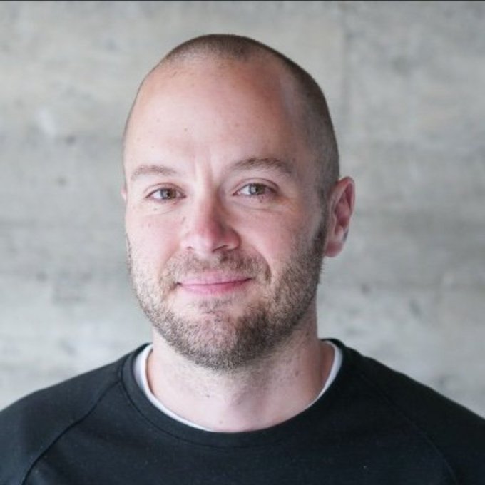 Headshot of a smiling bald man with a short beard wearing a black crew neck shirt against a gray concrete wall.