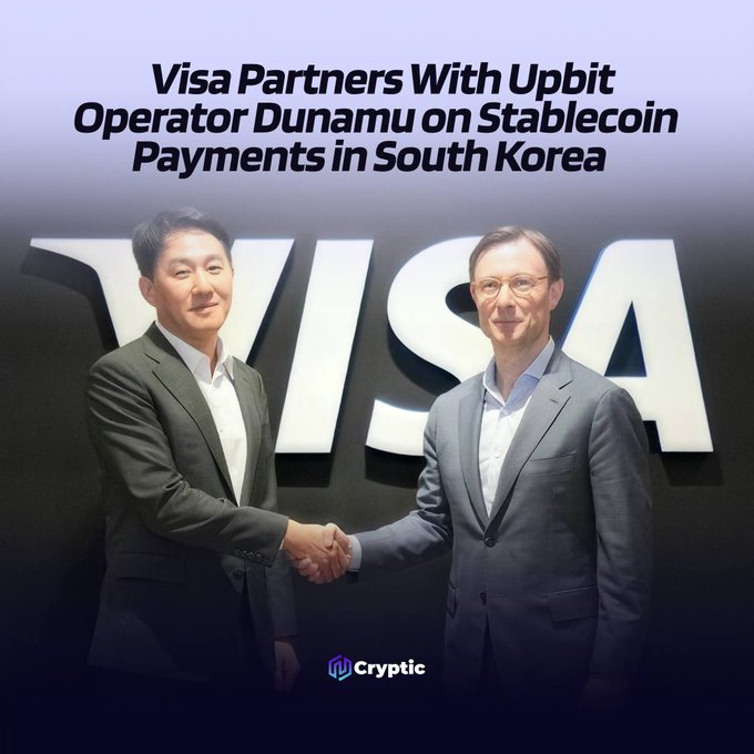 Two executives shake hands in front of a large Visa logo, illustrating the partnership between Visa and Dunamu—operator of South Korea's Upbit exchange—to collaborate on stablecoin payments, cross-border remittances, and AI-powered commerce.