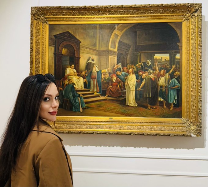 A woman with long dark hair and sunglasses on her head posing in an art gallery in front of a large, ornately gold-framed classical painting depicting a crowded religious or historical scene.