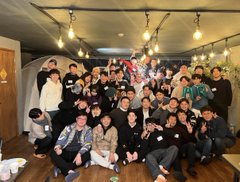 The image shows a large group of young men posing together for a photo. They are all dressed similarly, wearing black clothing that matches their smiles as they gather around each other. The room has a cozy atmosphere, with potted plants adding greenery to space. A tent can be seen in the background, suggesting an outdoor event or gathering. The post is written in Korean and includes a caption about happy 4th anniversary, which indicates that this group of friends might have gathered for a special occasion or milestone celebration.