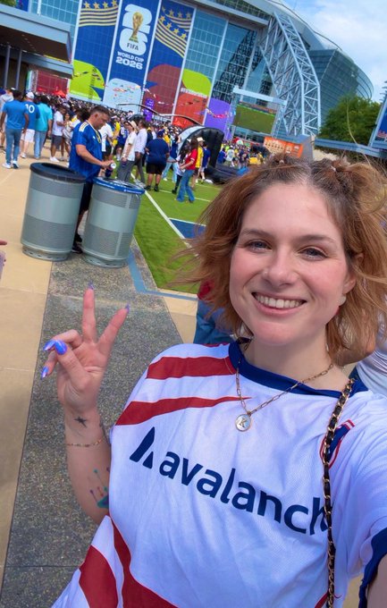 A selfie of a smiling woman with reddish-brown hair wearing an Avalanche jersey and peace sign, taken at a FIFA World Cup 2026 stadium with crowds and blue jerseys visible in the background.