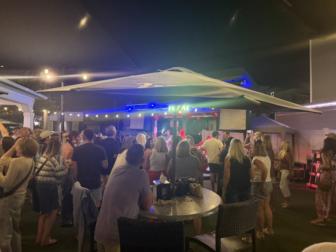 A nighttime outdoor scene at a venue called 'OZAR', showing a crowd of people gathered under a lit canopy watching a live performance on a stage illuminated with blue and red lights.