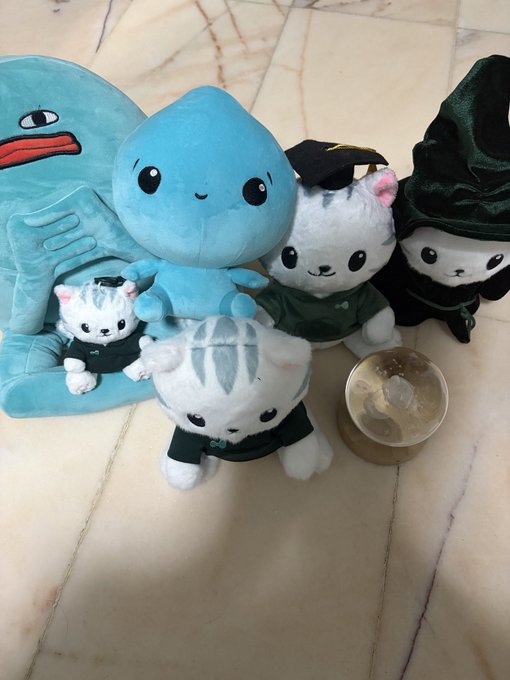 A collection of plush toys including a blue octopus-like character, a blue Squirtle-style plush, and three cat plushies wearing graduation caps and green sweaters, arranged on a marble floor next to a brass singing bowl.