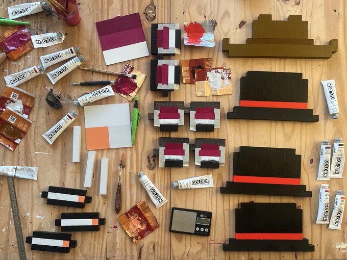 A wooden worktable scattered with art supplies including paint tubes, palette knives, small digital scales, and several small black, white, and red artworks in progress.