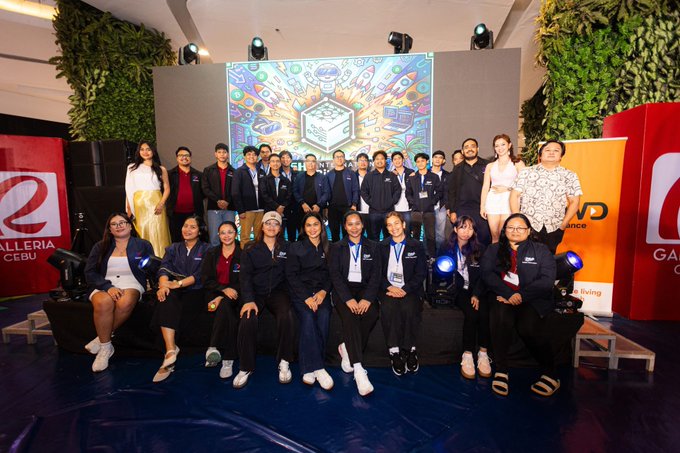 A group photo of young people in matching dark blue blazers posing on stage at a 'Galleria Cebu' event, with a colorful digital screen displaying tech-related graphics behind them.