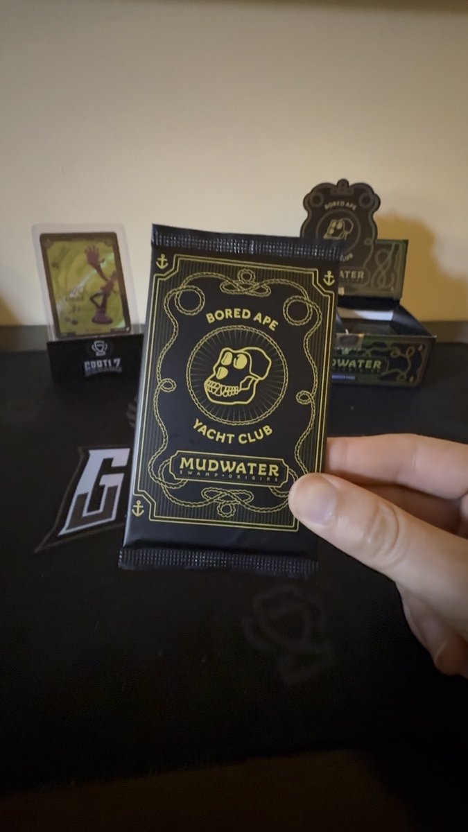 A hand displays a black snack package labeled “BORED APE YACHT CLUB MUDWATER,” with matching branded boxes and collectible-style packaging arranged behind it.