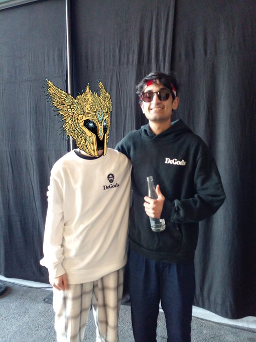 Two people pose together in front of a black backdrop; one wears a golden winged mask and a white "DeGods" long-sleeve, while the other wears sunglasses, a red headband, and a black "DeGods" hoodie while holding a water bottle.