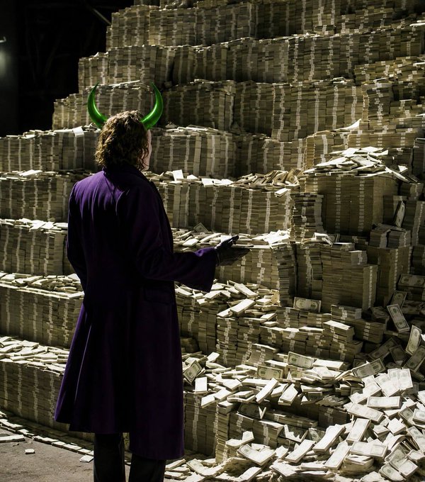 A person wearing a purple coat and green horn-like headwear stands in front of a massive wall of stacked cash, reaching toward the bills.