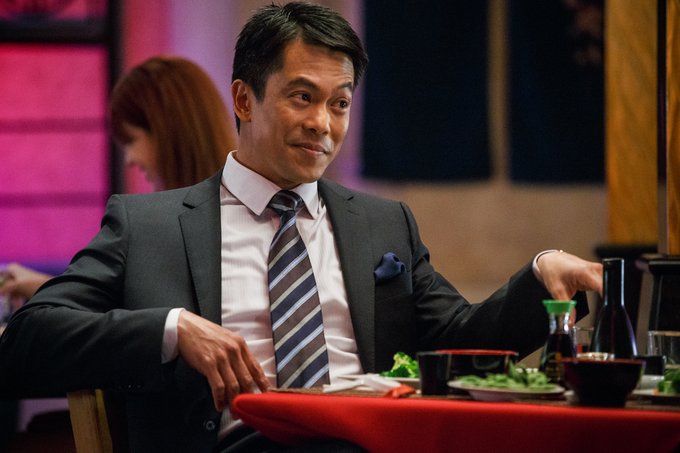 A sharply dressed man in a suit and striped tie sits at a dining table with sushi and sake, giving a knowing, sly look to the side—mirroring the post's smug tone about stacking low-vol leverage on top of meme-stock gambling to double-down on risk while pretending to play it safe.