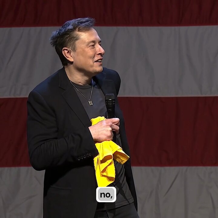 The image features a man standing on stage, holding a yellow towel or cloth over his shoulder while speaking into a microphone. The background of the image includes an American flag with red stripes, which adds context to the event taking place. The caption below the photo reads "I make dogecoin jokes and stuff because I kind of like dogecoin because it got the best sense of humor and it has dogs and memes and I love all those things." This suggests that the man is giving a humorous speech or presentation related to dogecoin, which he finds amusing.