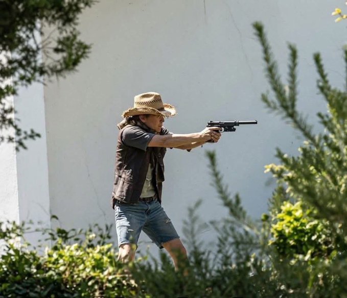 A man wearing a straw cowboy hat, vest, and denim shorts aiming a rifle while standing in front of a white wall, framed by green foliage.