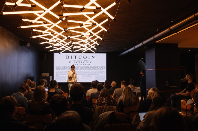 The image shows a large group of people gathered around a projector screen displaying information about Bitcoin. The audience appears engaged, with some individuals standing up and listening attentively while others are seated. The room has a dark color scheme, creating an atmosphere suitable for learning and discussion. A person is speaking into the microphone at the front of the room, likely providing more details or answering questions related to Bitcoin.
