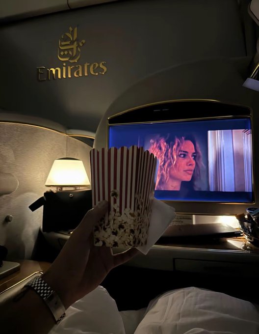A passenger enjoys Emirates first-class hospitality while watching a movie on the seat-back screen, holding up a striped popcorn box to capture the cozy in-flight entertainment experience—a luxurious way to "make some greens" on a good day.