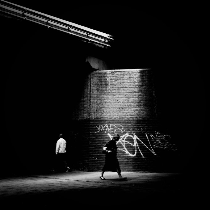 A black-and-white photograph captures a dimly lit urban scene where two figures walk past a brick section illuminated by overhead lighting, with graffiti visible on the wall.
