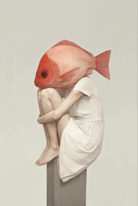 A melancholic figure in a white dress sits curled atop a concrete pillar with a large red fish head, embodying the surreal, introspective nature of the artwork "MELANCHOLIA" being displayed at the Time to Be Happy Gallery in New York.