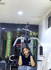 A young person smiles while posing in front of a home gym multi-station machine, wearing a dark shirt with a green plant visible in the background.