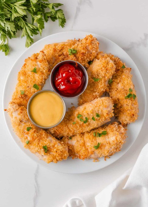 A white plate of crispy golden chicken tenders served with small bowls of ketchup and mustard sauce, garnished with parsley and fresh herbs on a marble surface.