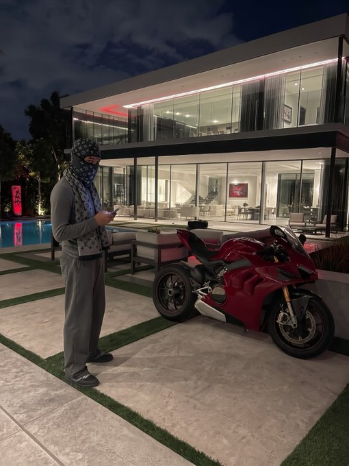 A masked person standing next to a red Ducati motorcycle in front of a modern luxury home with floor-to-ceiling windows and an illuminated pool area.