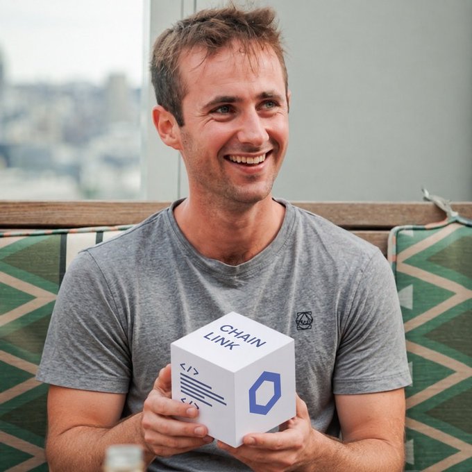 A smiling man in a gray t-shirt holds a white cube displaying the Chainlink logo and the text 'CHAIN LINK' with code-like symbols.