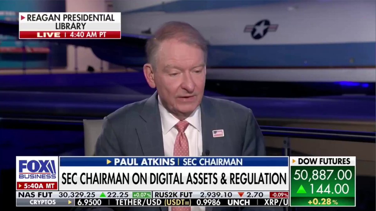 The image shows a man sitting at a desk, wearing a suit and tie, with his head down looking serious as he speaks into a microphone. The background features an airplane, suggesting that this scene takes place in an airport or near one. The text on the screen reads "SEC CHAIRMAN PAUL ATKINS just said the quiet part out loud! Where lawyers cannot give clients certainty on whether something is legal or not that creates NEEDLESS COST." This quote appears to be related to a recent speech given by the SEC Chairman, and it seems to be discussing the impact of uncertainty in the legal field.