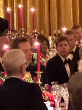 A dimly lit, elegant banquet scene showing formally dressed guests seated at tables adorned with pink taper candles and floral centerpieces.