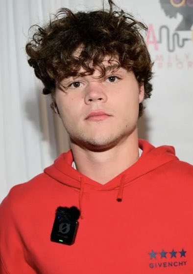 A young man with curly brown hair wears a red Givenchy hoodie featuring a wireless microphone clipped to the front. The post's reference to Jack Doherty buying $JACK daily connects to the man's identity as the content creator mentioned.