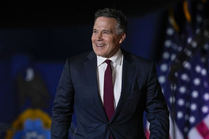 A man in a dark suit and burgundy tie smiles while walking on stage, with an American flag visible in the background.
