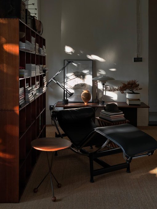A moody, atmospheric home office or reading nook featuring a black leather lounge chair with ottoman, a wooden bookshelf filled with books, a desk lamp casting dramatic light patterns on the wall, and decorative ceramics and plants.