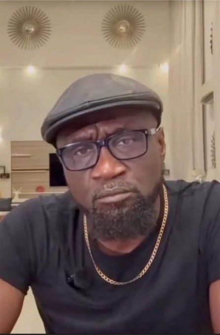 A selfie-style photo of a person with a beard wearing a gray flat cap, glasses, a black t-shirt, and a gold chain, in what appears to be an indoor setting with decorative gold wall art.