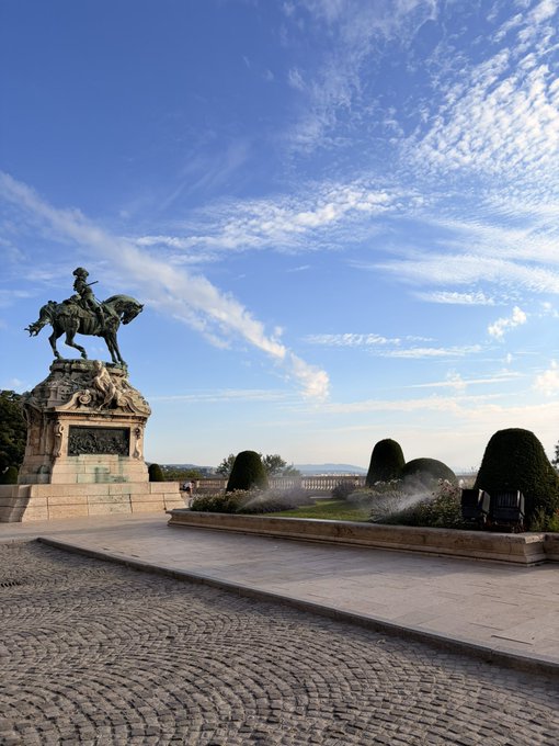 An equestrian statue of a historical figure on a stone pedestal stands in a plaza with manicured topiary gardens, set against a bright blue sky with wispy clouds.