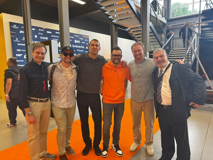 The image shows six men standing together, posing for a photo. They are all dressed in business attire, with one man wearing an orange shirt. The group appears to be gathered around a staircase or set of stairs within a building. In the background, there is a sign that reads "Too much powerlessness in a picture. You get a Physics of Bitcoin book if you guess right the full lineup." This suggests that they might have recently attended an event related to Bitcoin or cryptocurrency, and the photo captures their experience together.