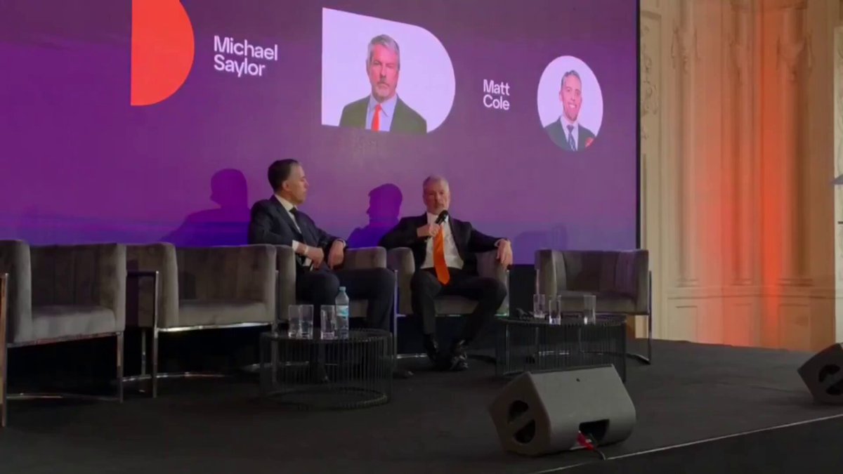 The image shows a stage with two men sitting next to each other, possibly participating in a panel discussion or conference. The stage has a purple background featuring three different faces of people, which could be representing the speakers at the event. There are also chairs placed around the stage for attendees. A microphone is visible on one side of the stage, indicating that it's being used by someone to speak into it during the event.