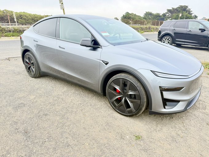 A silver Tesla Model Y Performance sits parked on a gravel lot, showcasing its sleek design, black performance wheels with red brake calipers, and Tesla's signature minimalist styling. The caption "Still enjoying slinging this guy around curving roads" reflects the owner's continued enjoyment of driving this high-performance EV, emphasizing its sporty handling capabilities.