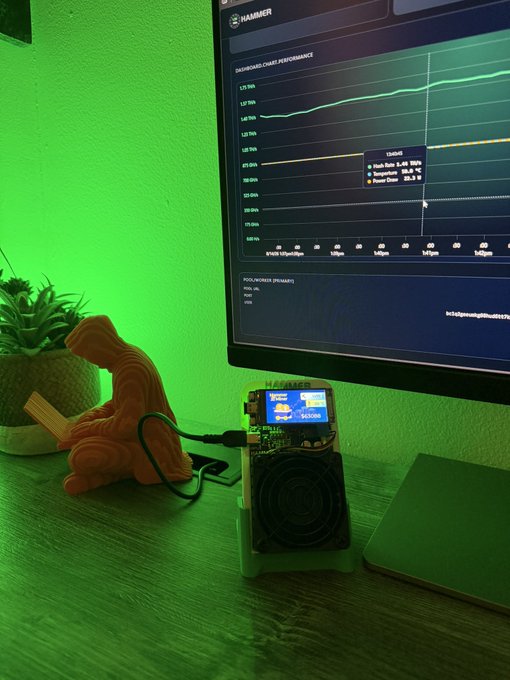 A desk setup illuminated by green lighting, featuring a small cryptocurrency mining device with a cooling fan and display screen running next to a computer monitor showing a hash rate graph, alongside a small Buddha statue and potted plant.