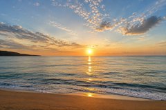 A serene landscape photograph of a sunset over the ocean, with the sun reflecting on the calm water and a sandy beach in the foreground.