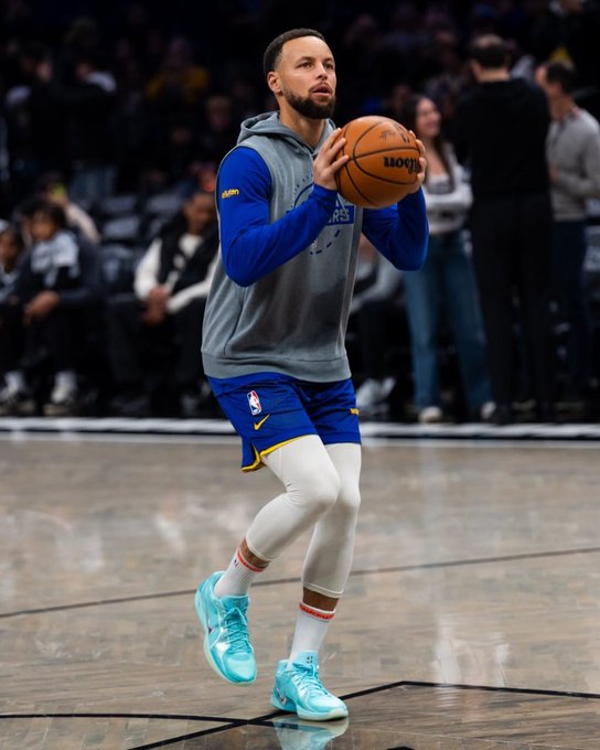 Steph Curry, wearing Golden State Warriors practice attire with light blue sneakers, is captured on a basketball court holding a Wilson basketball in a shooting stance.