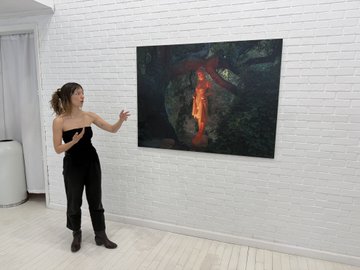 A woman in black clothing stands in a white-walled gallery, gesturing toward a large, dark abstract painting featuring red and black tones.