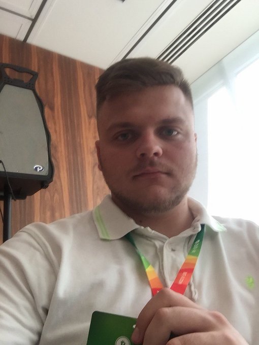 A selfie of a man wearing a white polo shirt with a red and yellow lanyard, holding a green badge.