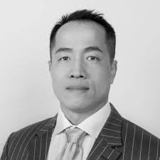 A black and white professional portrait shows an Asian man in a pinstripe suit and tie smiling against a light gray background.