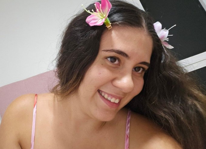 A smiling young woman with long dark hair wears two pink lily flowers in her hair and a pink top, posing for a selfie.