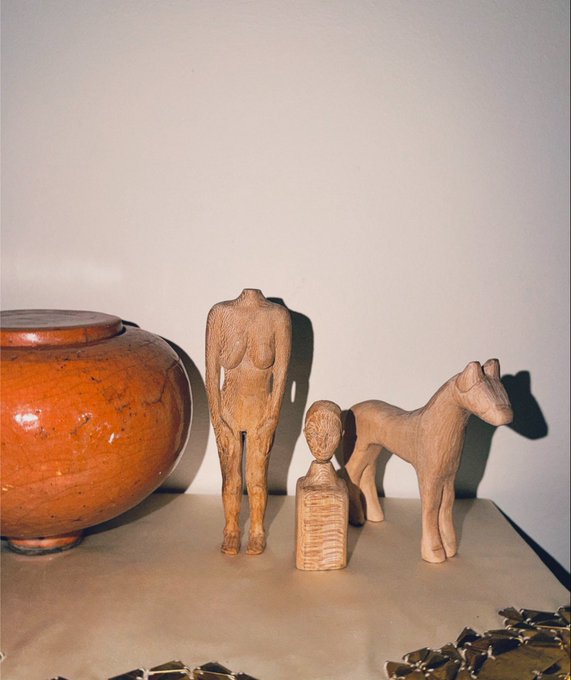 A still life of wooden carved folk-art figurines—a female figure, a small bust, and a horse—arranged on a surface next to a large terracotta pot.